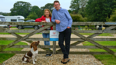 Man in blue shirt standing next to woman in red jacket in front of gate with their dog nearby